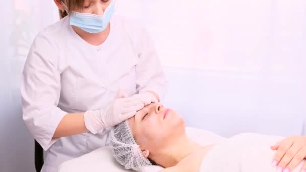 Cosmetólogo profesional en uniforme blanco, máscara médica y guantes aplica una máscara de rejuvenecimiento a la cara del paciente con movimientos de masaje de las manos. — Vídeos de Stock