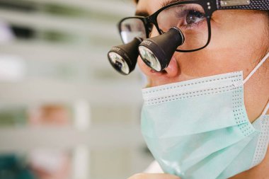 Diş hekimi muayenehanesinde dişçi loupe dürbünü ve tıbbi maske takan genç bir bayan doktor..