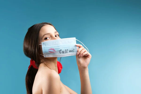 Een aantrekkelijk brunette meisje in het houden van de medische masker in een hand die haar gezicht met een kus teken en geschreven bel me. — Stockfoto