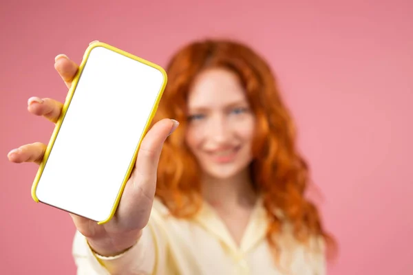 Rozmyte zdjęcie młodej damy z rudowłosą fryzurą i niebieskimi pięknymi oczami pokazującymi na aparacie telefon komórkowy z białym ekranem trzymającym w dłoniach na różowym tle. — Zdjęcie stockowe