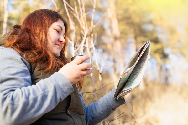 Foto vista lateral de uma mulher sorridente feliz ao ar livre com uma xícara de chá e um mapa segurando em suas mãos, desfrutando de dia ensolarado de inverno. — Fotografia de Stock