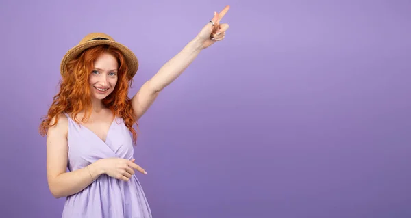 Bela jovem ruiva usando vestido de verão violeta e um chapéu posando para câmera segurando a mão apontando com um dedo. — Fotografia de Stock