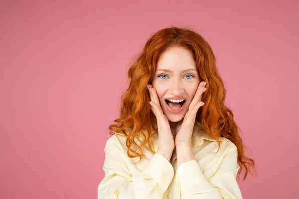 Menina bonita ruiva animado com olhos azuis e abriu a boca mantendo as palmas das mãos nas bochechas posando sobre um fundo rosa. — Fotografia de Stock