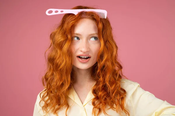 Image de belle fille rousse avec un peigne à cheveux fins sur sa tête posant pour la caméra sur fond rose, concept de cheveux coupés. — Photo