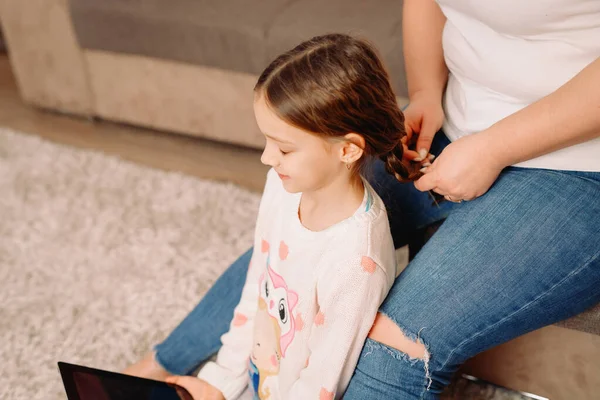 Foto recortada de mãe em jeans sentada no sofá e tecendo tranças para sua filha em casa, menina sentada com os olhos fechados. — Fotografia de Stock