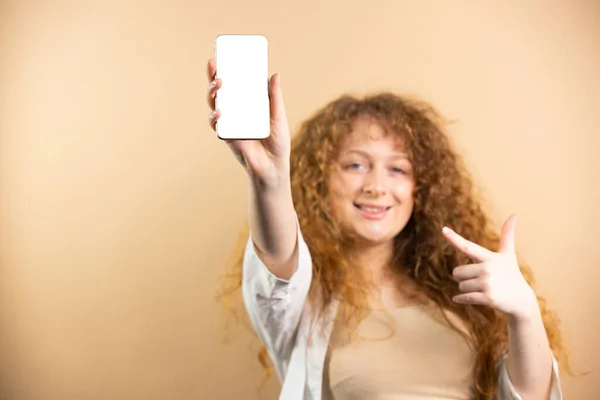 Belle femme modèle avec coiffure bouclée levant la main vers le haut montrant le smartphone avec écran blanc vide sur fond doré. — Photo