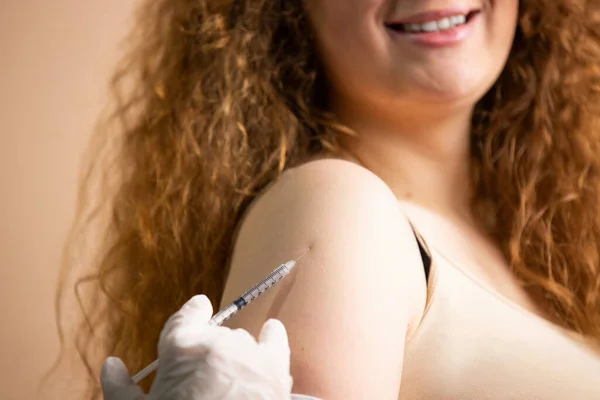 Mujer joven recortada con el pelo rojo sonriendo ampliamente vacunarse con la vacuna contra el coronavirus, médico en guantes estériles usando seringe para inyección. — Foto de Stock