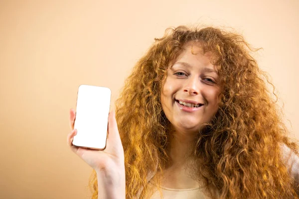 Beauty redhead girl with long and shiny wavy red hairsmiling widely holding in her hand smartphone with white screen over golden background. — Stock Photo, Image