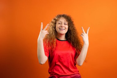 Futbol takımının kırmızı üniformalı, neşeli, kızıl saçlı, gülümseyen kadını zafer işareti olarak parmaklarını kaldırıyor..