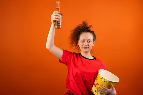 Uvolněný fanouškovský fotbal žena v červené uniformě s miskou popcornu a udržet láhev piva zvedající ruku nahoru. — Stock fotografie
