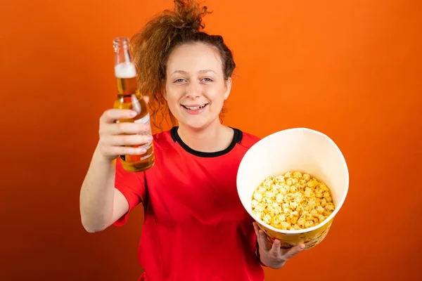 Leende kvinna med ponny svans hejar och stödja fotbollslag som håller en skål popcorn och flaska kall öl firar seger. — Stockfoto