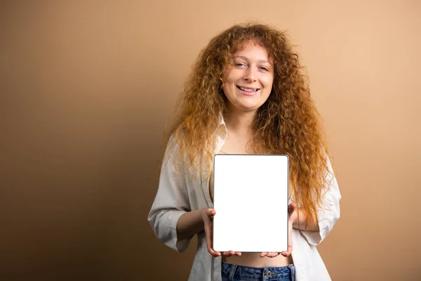 Счастливая веселая рыжая молодая женщина с улыбкой лица, держа перед планшетом с белым экраном на золотом фоне. — стоковое фото