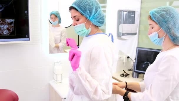 En assistent i mask hjälpa tandläkare kvinna sätta på uniform i skåp med röntgen foto på monitorer för att börja arbeta. — Stockvideo