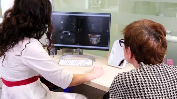 Врач-стоматолог в медицинской маске сидит за столом и показывает рентген зубов на компьютере перед пациентом в стоматологической клинике. — стоковое видео