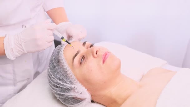Спокойная и расслабленная женщина в медицинской шапочке лежит на массажном столе и получает инъекцию красоты, сделанную косметологом в салоне красоты. Не поддающаяся инъекциям процедура — стоковое видео