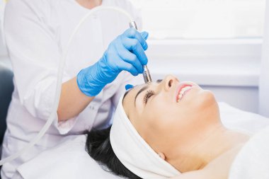 Güzellik uzmanı ve mutlu bir kadının kesilmiş fotoğrafı masaj masasında uzanıyor ve bir kozmetik kliniğinde mavi eldivenli bir güzellik uzmanı tarafından mikro dermabrüksiyon yaptırılıyor..