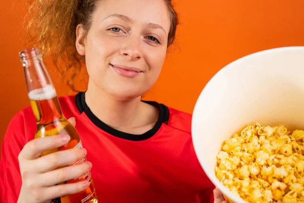 Beskuren bild av en rödhårig kvinna fan av fotboll i röd t-shirt poserar med en flaska öl och en skål popcorn. — Stockfoto