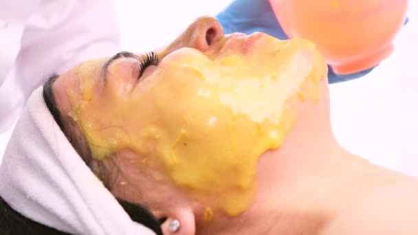 Professionell kosmetolog gör procedurer för klient i ljusskåp tillämpa gul ansiktsföryngring kamomill alginat mask. — Stockvideo
