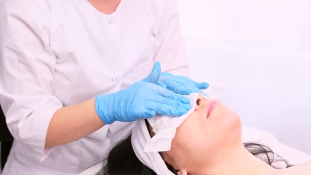 Cosmetologist em luvas azuis removendo a umidade do rosto da mulher usando guardanapo de papel descartável no salão de spa, conceito de medicina alternativa. — Vídeo de Stock