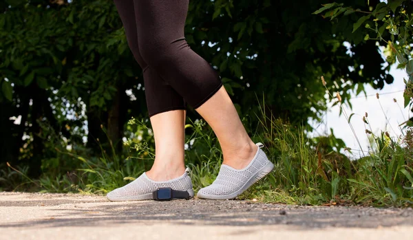 Photo recadrée de jambes de femme dans des leggings et des chaussures confortables debout près de la forêt sur la route et la montre numérique est couché sur le sol. — Photo