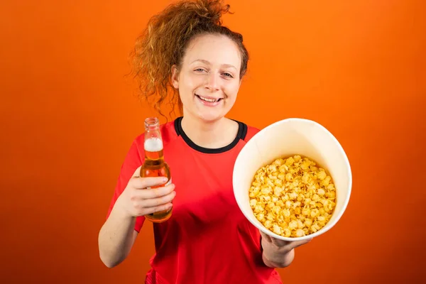 Leende ung kvinna fotboll fan titta match stöd favorit lag bär team uniform hålla en hink popcorn och alkohol dryck. — Stockfoto
