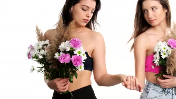 Unga vackra attraktiva blonda och brunett flickor som håller händer tittar på varandra med ömhet och täcker sitt bröst med blommor. — Stockvideo
