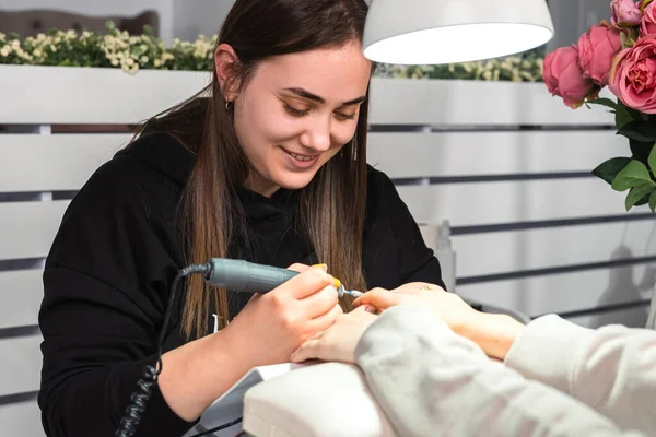 Master professional in schoonheid salon in zwart uniform met behulp van elektrische nagel bestand boor maken manicure aan een vrouw klant. — Stockfoto