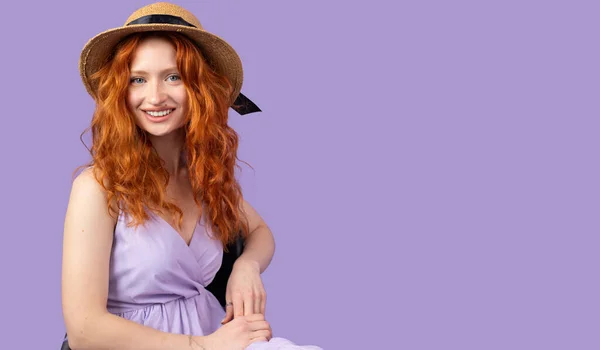 Photo d'une belle rousse jeune femme souriante portant largement robe de soleil et chapeau de paille pourl'été assis sur la chaise sur fond violet et l'espace de copie. — Photo