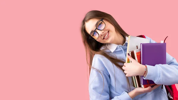 Vtipné usměvavé školní dívka s batohem a knihy v rukou v uniformě s brýlemi vypadá šťastně na růžovém pozadí a kopírovat prostor. — Stock fotografie