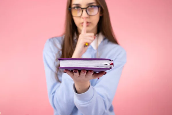 Foto borrada de uma menina bonita estudante vestindo óculos em uniforme fazendo sinal de silêncio com um figer índice e segurando um livro em sua mão. — Fotografia de Stock