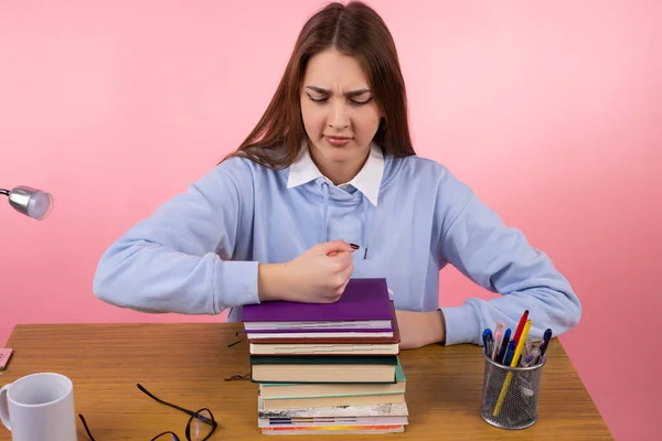 Krásná blond naštvaný mladý student sedí u stolu v vysoké škole modré uniformě dal její pěst na hromadu knih na růžovém pozadí. — Stock fotografie