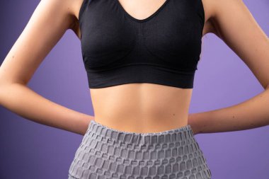 İnce vücutlu, siyah bluzlu, ellerini arkada tutarak mor arka planda poz veren genç bir kadının kesilmiş fotoğrafı..