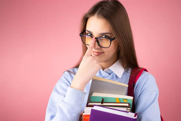 Jovem estudante universitária loira atraente segurando livros em suas mãos com mochila e corrige seus óculos posando em fundo rosa. — Fotografia de Stock