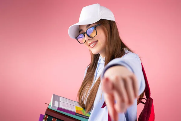 Legal bonita loira escola menina sorrindo vestindo óculos e um boné segurando livros e apontando com um dedo para a câmera. — Fotografia de Stock