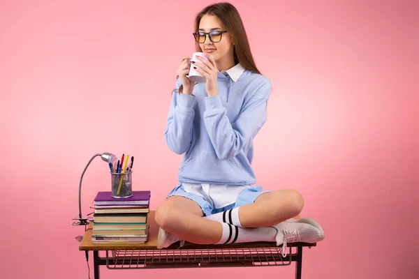 Linda menina estudante em óculos e uniforme sentado na mesa com livros tendo uma pausa beber uma xícara de chá. — Fotografia de Stock