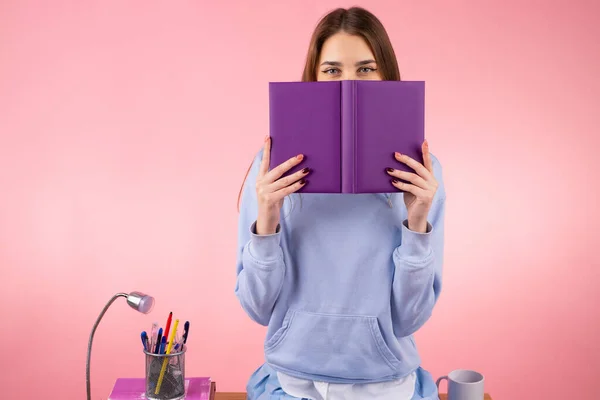 Imagem de uma estudante linda menina com cabelo loiro cobrindo seu rosto com um livro de cor violeta e sentado na mesa. — Fotografia de Stock