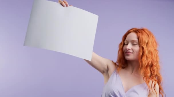 Mycket skrattande rödhårig ung kvinna håller upp vitt papper i handen och använder pekfingret på en annan hand pekar på kopia utrymme för reklam. — Stockvideo