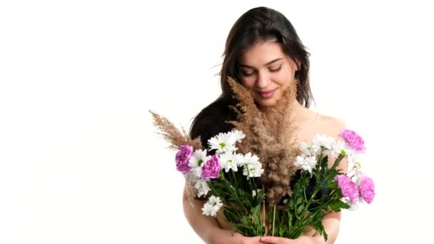 Ενιαία νεαρή όμορφη κοπέλα μελαχρινή χαμογελά καλύπτει γυμνό στήθος της με λουλούδια και τα μαλλιά της φυσάει στον άνεμο πάνω από το λευκό φόντο. — Αρχείο Βίντεο