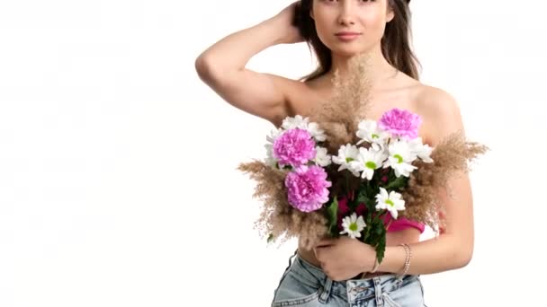 Nuori kaunis nainen tilalla kukkakimppu kattaa hänen alasti rintojen hiukset puhaltavat tuulessa valkoisella taustalla. — kuvapankkivideo