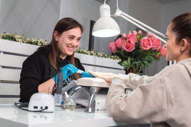 Eldiven takan genç manikürcü, güzellik salonunda manikür yapan genç bir kadının eski manikürünü çıkarırken müşterisine bakıyor..