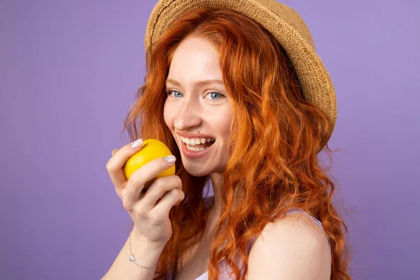 Молода руда жінка з блакитними очима в солом'яному капелюсі, що посміхається, широко тримає яблуко, готове їсти на фіолетовому фоні . — стокове фото
