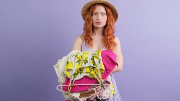 Serious young redhead woman in a hat looking at the camera and changing her facial emotion in a happy face while looking at the clothes she holding in her hands. — Stock Video