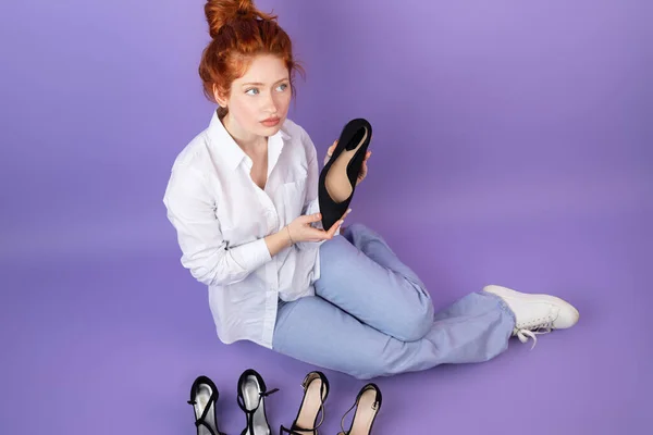 Nachdenkliches hübsches junges Mädchen mit roten Haaren in lässiger Kleidung mit einem Schuh in der Hand auf dem Boden sitzend, welches Paar Schuhe es zu kaufen gibt. — Stockfoto