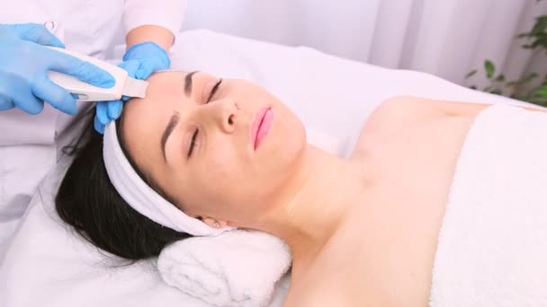 De arts-cosmetoloog maakt ultrasone reiniging procedure van de gezichtshuid van een mooie jonge vrouw in een schoonheidssalon. — Stockvideo