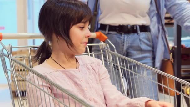 Egy fiatal nő, aki két almát ad a lányának a bevásárlókocsiban ülve, és a lány csukott szemmel szagolja őket, gyümölcsöt vesz a szupermarketben.. — Stock videók