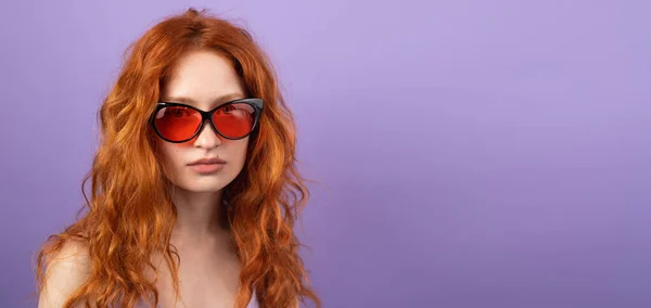 Jolie jeune femme aux cheveux roux portant des lunettes de soleil pour l'heure d'été posant sur fond violet avec espace de copie. — Photo