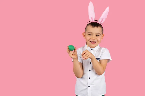 Фото усміхненого чарівного хлопчика з вухами кролика на голові і білою футболкою, що тримає зелене яйце і пензлем для малювання в руках позує на рожевому тлі з копією простору . — стокове фото