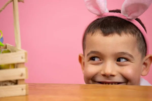 Glad liten pojke med kanin öron på huvudet tittar från under bordet med sluga ögon i korgen på rosa bakgrund. — Stockfoto