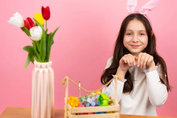 Söt liten flicka bär kanin öron på huvudet vikta händerna som kanin sitter vid bordet med korg och vas med blommor och tittar på kameran. — Stockfoto