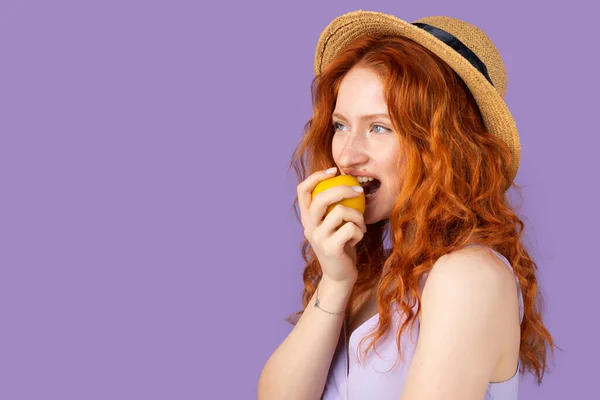 Портрет молодої красивої привабливої дівчини з рудим волоссям модний капелюх кусає жовте яблуко на фіолетовому фоні та копіює простір . — стокове фото